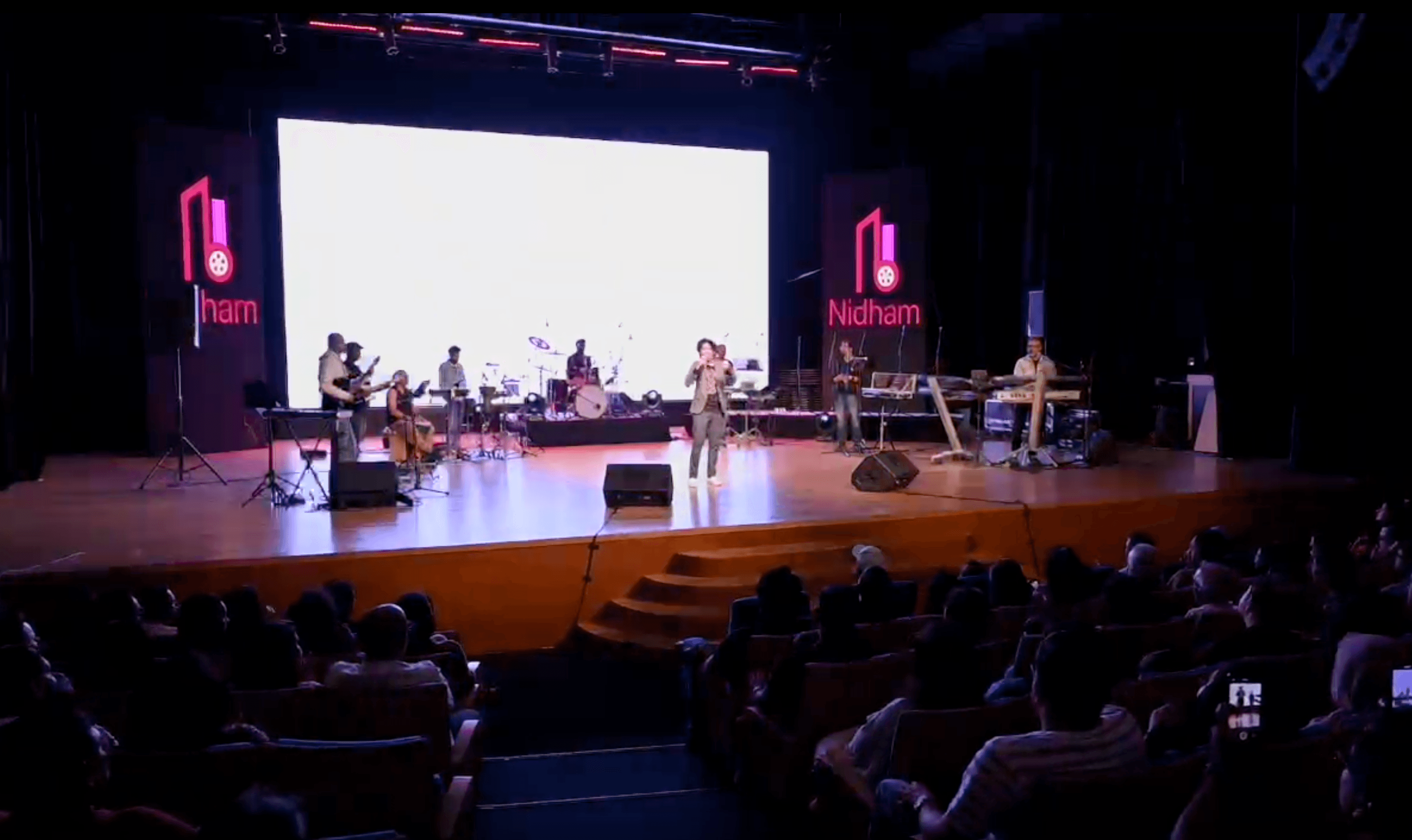 Live band and vocalist performing on stage under a Nidham-branded backdrop, audience seated in the foreground.